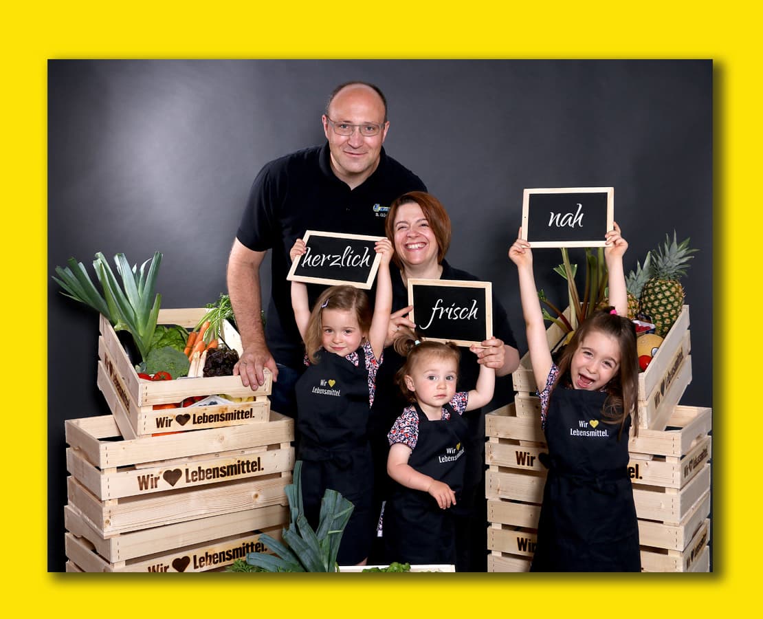 Familie Weinle Ein fröhliches Foto der Familie Weinle zwischen Obst- und Gemüsekisten auf gelbem Hintergrund