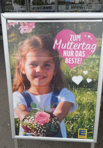 Muttertagsplakat von EDEKA mit lächelndem Mädchen und Blumenstrauß vor Frühlingswiese sowie dem Schriftzug „Zum Muttertag nur das Beste!“.