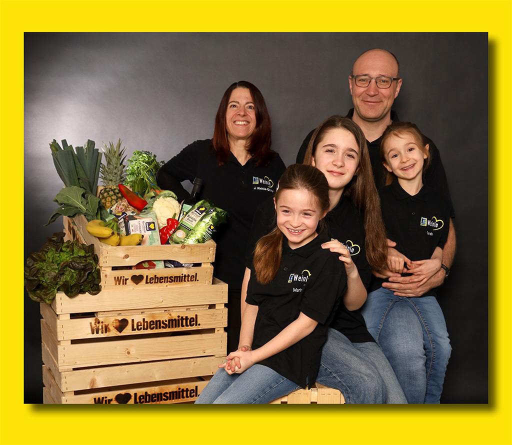 Das Ehepaar Weile-Günter mit ihren 3 Kindern lachend neben gefüllten Einkaufskisten mit frischer Ware und der Aufschrift "Wie lieben Lebensmittel"
